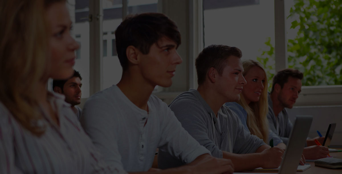 People sit in a classroom, attentively watching a presentation, with open laptops and notebooks in front of them.